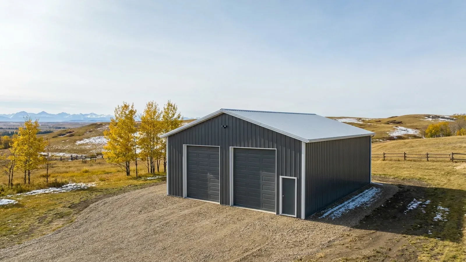 A steel shop with two overhead doors and a man door on a gravel pad, aspen trees and rolling foothills behind