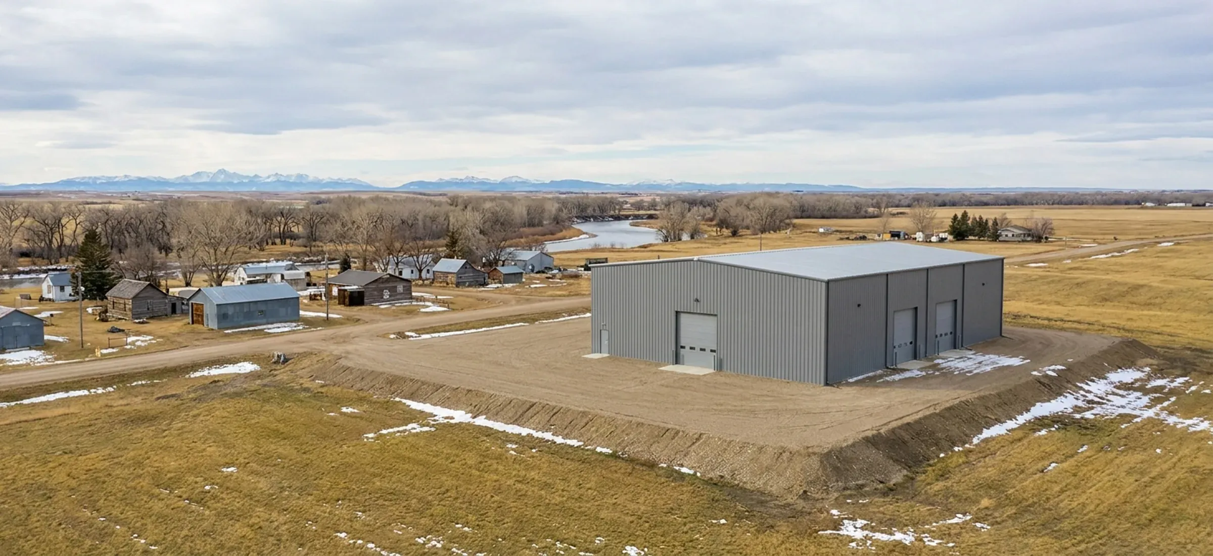 A steel industrial building on a raised gravel pad near a river valley with mountains on the horizon
