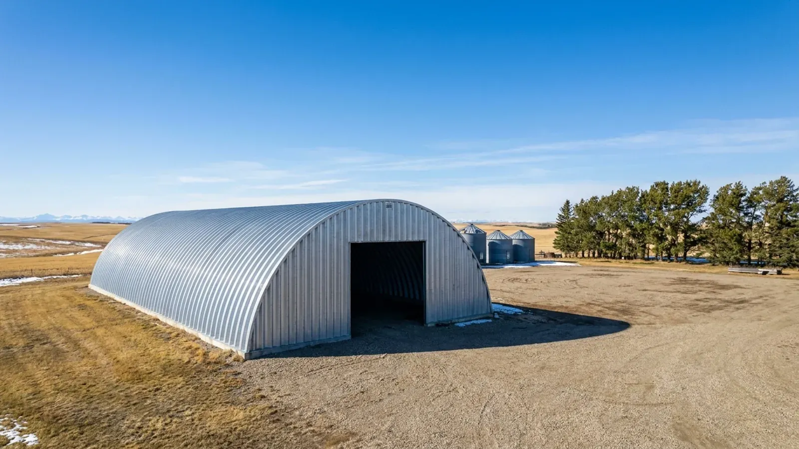 A galvanized arch-style quonset storage building with an open end wall on a prairie farmyard